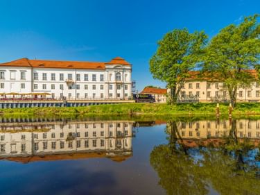 Schloss Oranienburg, ein weißes Barockgebäude mit orangefarbenem Dach, spiegelt sich im ruhigen Wasser eines Flusses. Grüne Bäume rahmen die Szene.