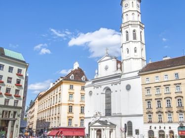 Pferdekutschen auf Kopfsteinpflasterplatz vor weißer Kirche mit hohem Turm. Historische Gebäude umgeben den Platz unter blauem Himmel.