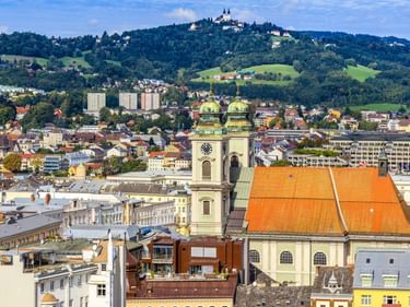 Luftaufnahme der Linzer Altstadt mit Kirche mit orangefarbenem Dach und zwei grünen Kuppeln. Pöstlingberg mit Wallfahrtskirche im Hintergrund.