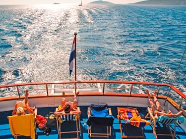 Sonnendeck der MS Columbo mit bunten Liegestühlen und kroatischer Flagge. Blaues Meer mit Kielwasser, Segelboot und Küstenhügeln im Hintergrund.