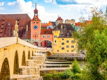 Steinerne Brücke in Regensburg mit mittelalterlichen Bögen über die Donau. Farbenfrohe historische Gebäude und ein Uhrturm in der Altstadt.
