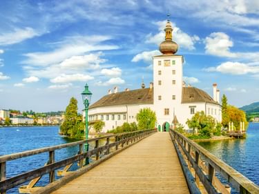 Holzsteg führt zu Schloss Orth, einem weißen Schloss mit Turm auf einer kleinen Insel im Traunsee bei Gmunden, Österreich.