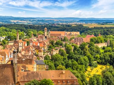 Luftaufnahme von Rothenburg mit mittelalterlichen Gebäuden, roten Ziegeldächern, Kirchtürmen und grünen Wäldern. Hügelige Landschaft im Hintergrund.