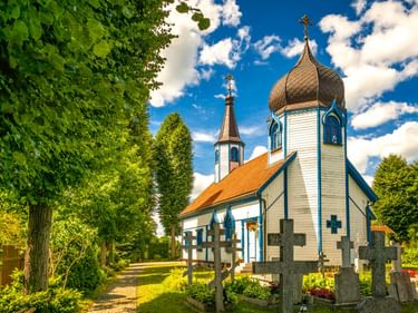 Weiße Holzkirche mit Zwiebeldach und orangefarbenem Dach in Wojnowo, umgeben von grünen Bäumen und Friedhofskreuzen unter blauem Himmel.