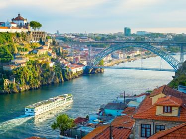 Flusskreuzfahrtschiff auf dem Douro in Porto mit ikonischer Metallbogenbrücke, historischen Gebäuden am Hang und roten Ziegeldächern im Vordergrund.