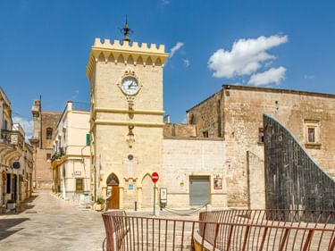 Historischer Uhrturm mit Zinnen auf dem Stadtplatz von Avetrana, umgeben von traditionellen Steingebäuden unter blauem Himmel.