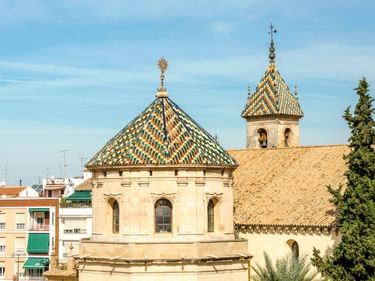 Historische Kirche in Lucena mit zwei bunten Kuppeln und einem Glockenturm. Das Gebäude hat cremefarbene Wände und dekorative Dächer.