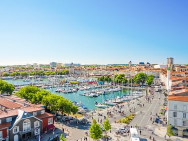 Luftaufnahme des Hafens von La Rochelle mit zahlreichen Segelbooten, Uferpromenade und historischer Altstadt mit roten Ziegeldächern unter blauem Himmel.