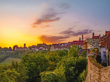 Sonnenuntergang über Rothenburgs mittelalterlicher Altstadt mit roten Dächern, Kirchtürmen und historischen Gebäuden über grünen Weinbergen.