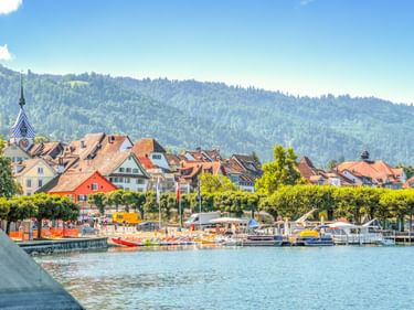 Uferpromenade in Zug mit bunten historischen Gebäuden, Kirchturm, Hafen mit Booten und bewaldeten Bergen im Hintergrund.