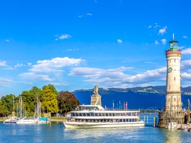 Lindauer Hafen am Bodensee mit weißem Leuchtturm, Löwenstatue, Fahrgastschiff, Segelbooten und Bergen im Hintergrund unter blauem Himmel.