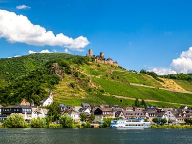 Alken an der Mosel mit Burg Thurant auf einem Hügel. Weinberge bedecken die Hänge, Häuser säumen das Flussufer, ein Boot liegt vor Anker.