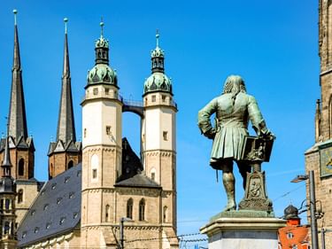 Bronzestatue vor historischen Kirchtürmen in Lutherstadt Wittenberg unter klarem blauen Himmel. Zwillingstürme und Kuppeltürme sichtbar.