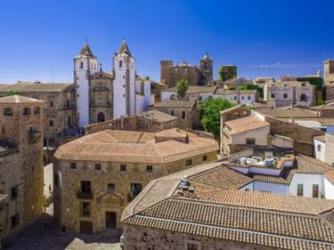 Luftaufnahme der Altstadt von Cáceres mit Terrakotta-Dächern, weißer Kirche mit Zwillingstürmen und mittelalterlichen Steingebäuden.