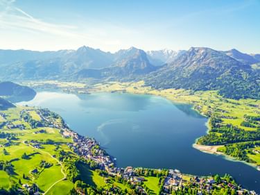 Luftaufnahme vom Wolfgangsee umgeben von grünen Wiesen und Bergen. Kleine Dörfer säumen die Ufer, mit Alpengipfeln im Hintergrund.