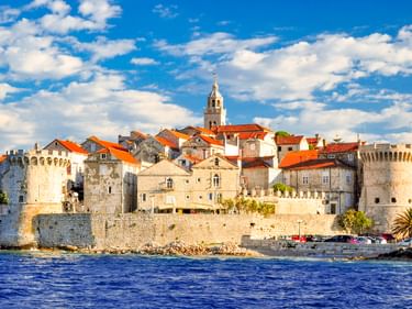 Altstadt Korcula mit Steinmauern, Türmen und roten Ziegeldächern vom Meer aus gesehen. Ein Kirchturm erhebt sich über den mittelalterlichen Gebäuden unter blauem Himmel.