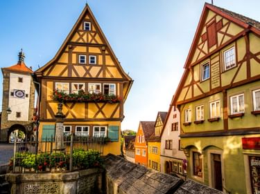 Bunte Fachwerkhäuser in Rothenburg mit einem Brunnen im Vordergrund und einem Torturm links unter blauem Himmel.