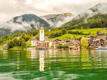 St. Wolfgang mit weißem Kirchturm am grünen Seeufer, traditionelle Alpengebäude, bewaldete Berge mit Nebel im Hintergrund.