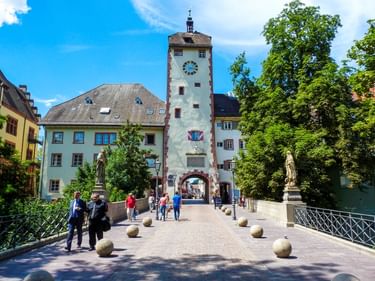 Weißer Turm mit Uhr und Torbogen in Waldshut, flankiert von bunten Häusern. Fußgänger auf Brücke mit Steinkugeln und Statuen.