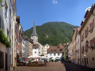 Fußgängerplatz in Churs Altstadt mit bunten Gebäuden, Kirchturm, Straßencafés und grünem Bergpanorama unter blauem Himmel.