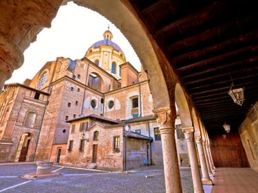 Blick durch Arkadenbogen auf historische Backsteinkirche mit blauer Kuppel an der Piazza delle Erbe in Mantua, Italien. Holzbalkendecke sichtbar.