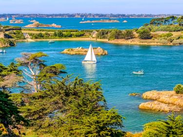 Weißes Segelboot auf türkisfarbenem Wasser nahe felsigen Inseln mit Pinien auf der Île de Bréhat. Steinhäuser und grüne Vegetation am Ufer.