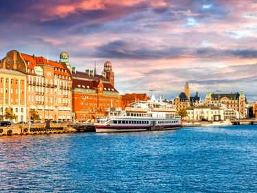 Farbenfrohe historische Gebäude entlang der Uferpromenade von Malmö mit einer Personenfähre am Hafen unter dramatischem Wolkenhimmel.