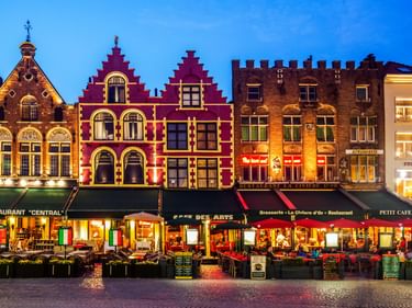 Historische flämische Giebelhäuser am Grote Markt in der Abenddämmerung beleuchtet. Bunte Fassaden mit Restaurants und Außensitzplätzen auf Kopfsteinpflaster.