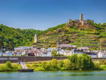 Kobern-Gondorf an der Mosel mit terrassierten Weinbergen am Hang und Burgruine auf dem Gipfel. Kirchtürme zwischen den Häusern sichtbar.