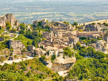 Panoramablick auf Les Baux, ein mittelalterliches Bergdorf mit Steingebäuden auf Felsenklippen, umgeben von grüner Vegetation und Ebenen.