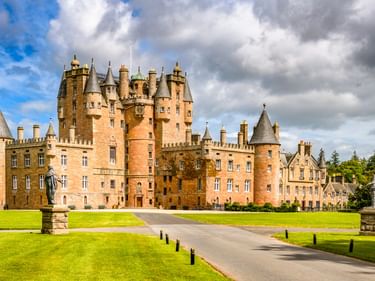 Glamis Castle in Angus mit mehreren Türmchen und Türmen aus rosa Sandstein, umgeben von grünen Rasenflächen unter dramatischem Wolkenhimmel.