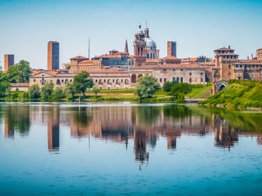 Historische Skyline von Mantua, Italien mit Domkuppel und Türmen, die sich im ruhigen Fluss Mincio spiegeln. Grüne Bäume säumen das Ufer.