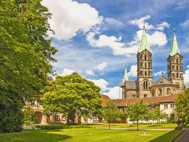 Bamberger Dom mit vier Türmen und grünen Kupferdächern, umgeben von historischen Gebäuden und einer grünen Rasenfläche mit Bäumen unter blauem Himmel.