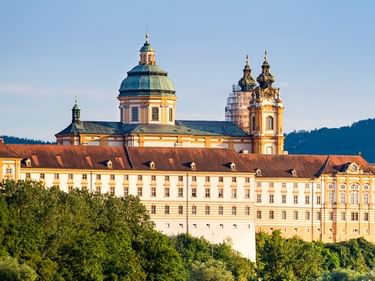 Stift Melk in Österreich, ein barockes Kloster mit gelber Fassade und Zwillingstürmen, auf einem Hügel über grünen Bäumen unter blauem Himmel.