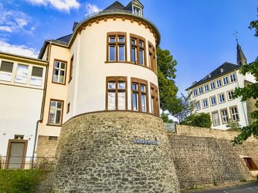 Alte römische Steinmauer in Bitburg mit cremefarbenem Gebäude mit rundem Turm und braunen Fenstern darüber, unter blauem Himmel.