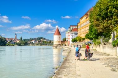 Radfahrer auf Uferweg entlang des Inn in Passau mit historischem Turm, bunten Häusern und Kirchturm unter blauem Himmel.