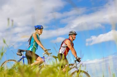 Zwei Radfahrer mit Helmen und Rucksäcken fahren mit Mountainbikes durch hohes Gras unter blauem Himmel mit weißen Wolken.