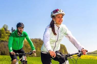 Zwei Radfahrer mit Helmen fahren durch grüne Landschaft. Frau in weißer Jacke führt, Mann in grüner Jacke folgt an sonnigem Tag.