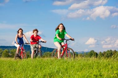Drei Kinder fahren auf roten Fahrrädern durch eine saftig grüne Wiese unter blauem Himmel mit weißen Wolken. Bewaldete Hügel im Hintergrund.
