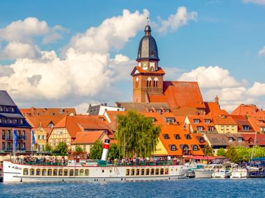 Hafenansicht von Waren Müritz mit Fahrgastschiff, traditionellen Gebäuden mit orangefarbenen Dächern und markanter Kirche unter blauem Himmel.