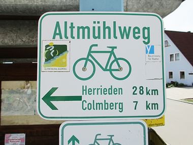 Weißes Wegweiserschild für den Altmühlweg mit Fahrradsymbol und Entfernungen: Herrieden 28km, Colmberg 7km. Informationstafel im Hintergrund.