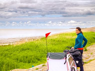 Frau in blauer Jacke mit Fahrrad und weißem Kinderanhänger mit roter Fahne auf Küstenweg in Ostfriesland. Grüne Dünen und Meer unter bewölktem Himmel.