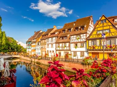 Traditionelle Fachwerkhäuser entlang eines Kanals im Klein-Venedig-Viertel von Colmar, mit leuchtenden Blumen im Vordergrund und blauem Himmel.