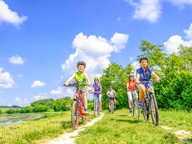 Fünf Radfahrer mit Helmen fahren auf einem Grasweg neben einem Fluss unter blauem Himmel mit weißen Wolken. Grüne Bäume und Hügel im Hintergrund.