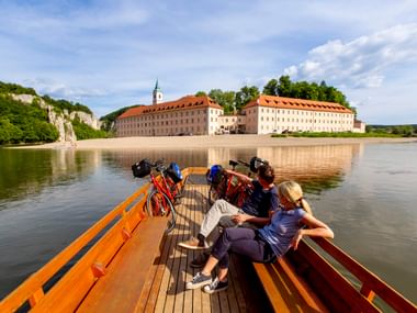 Radfahrer mit Rädern auf Holzboot auf der Altmühl vor Kloster Weltenburg, einer großen Klosteranlage mit orangefarbenen Dächern am Wasser.