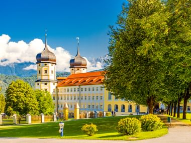 Stift Stams in Tirol mit gelbem Barockgebäude, zwei Zwiebeltürmen, orangem Dach, grüner Wiese und Baumallee unter blauem Himmel.