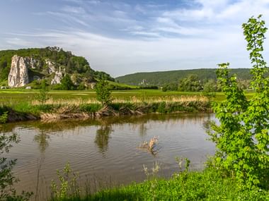 Ruhige Altmühl fließt durch grüne Wiesen mit weißen Kalksteinfelsen und bewaldeten Hügeln im Hintergrund unter blauem Himmel.