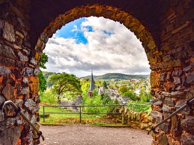 Blick durch einen verwitterten Steinbogen auf Saarburg mit Kirchturm, grünen Bäumen und sanften Hügeln unter bewölktem Himmel.