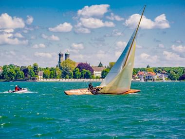 Holzsegelboot mit weißem Segel auf dem Bodensee bei Friedrichshafen. Kirchtürme und Gebäude am Ufer unter blauem Himmel sichtbar.