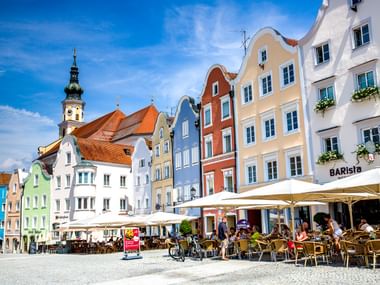 Historischer Stadtplatz in Schärding mit bunten Barockgebäuden, Straßencafés und Kirchturm unter blauem Himmel.